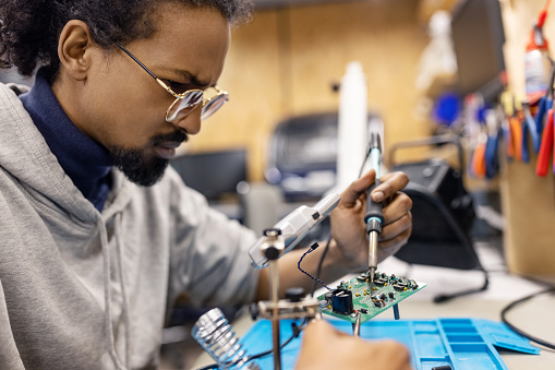 Male electrical engineer soldering electronics circuit board with a soldering iron at workshop. Young man preparing circuit board at makers space.