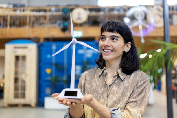 Smiling woman engineer standing in office holding windmill model Smiling woman engineer standing in office holding windmill model. Female professional working on sustainable energy at creative makers space. industrial designer stock pictures, royalty-free photos & images