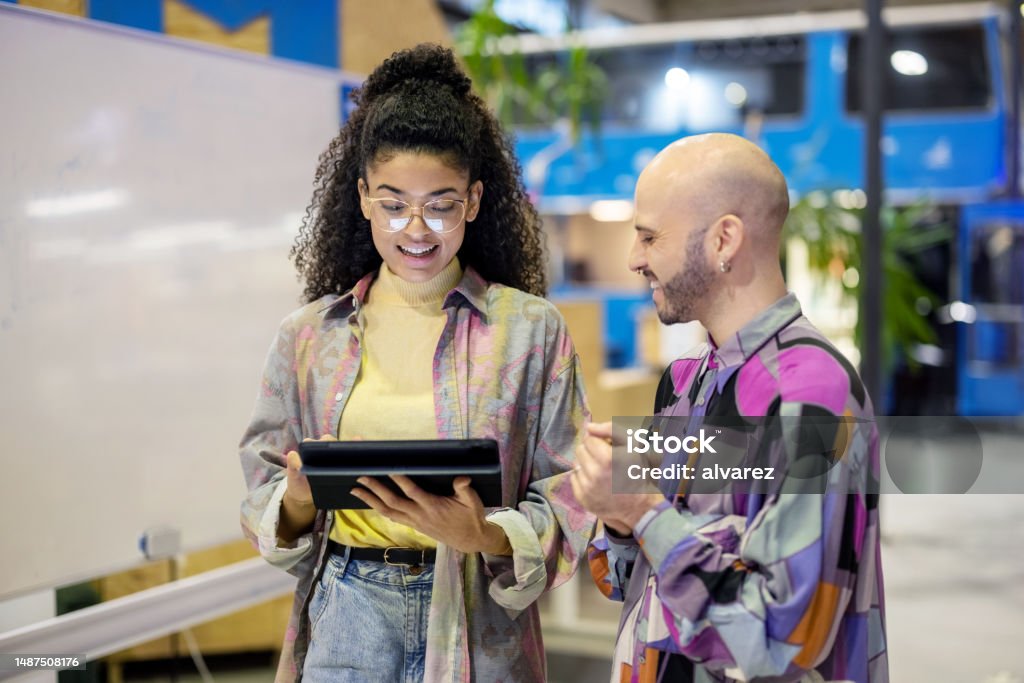 Two creative people walking together and discussing a project on a digital tablet at makers space Two young people walking together and discussing a project on a digital tablet at a creative workspace. Multiracial man and woman walking through maker space corridor and talking. Adult Stock Photo