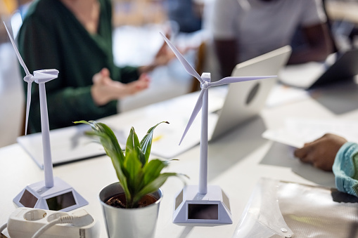 3D models of wind turbines on desk with design experts brainstorm ideas for sustainable wind energy solutions. Team of engineers having a meeting in an office to discuss a wind energy project.