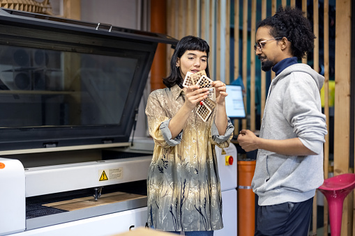Woman engineer holding laser cut wooden pieces and talking with male colleague at makers space. Two engineers working on wood laser cutting project at makers space.