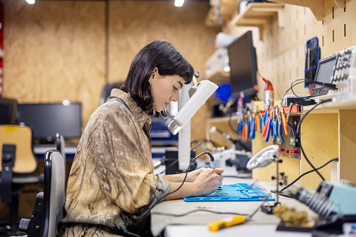 Side view of a woman preparing circuit boards at makers space. Female electrical engineer soldering electronics circuit board with a soldering iron at workshop.