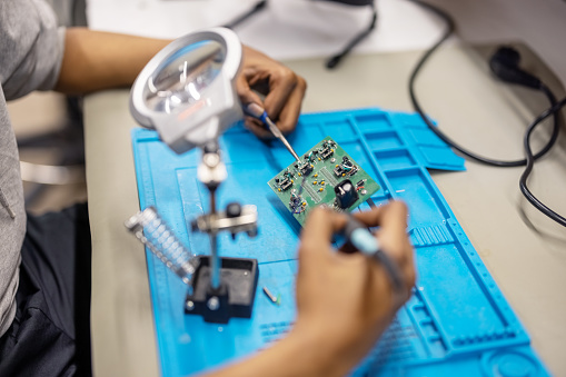 Close-up of a man welding circuit board under magnifying glass in workshop. Male electrical engineer soldering electronics circuit board with a soldering iron at workshop.