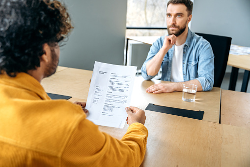 Human resources recruitment concept. Hiring interview between hr manager and hiring for job caucasian man, they sitting in the office, hr manager holds resume of job applicant, asks questions