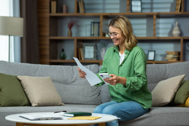 uma idosa recebeu boas notícias, acordo financeiro, pagamento de empréstimo, venda de casa, pensão, subsídio. ele está sentado no sofá de casa, segurando o telefone e documentos nas mãos. contas correntes - internet e mail paying credit card - fotografias e filmes do acervo