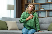 Heart attack, panic attack,. An elderly woman sits on the sofa at home and holds her hand to her chest. It is difficult to breathe, feels severe pain