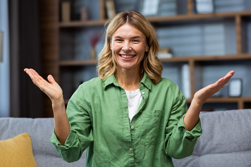 Portrait of a scared happy curvy woman sitting at home on the sofa and smilingly spreading her hands looking at the camera.