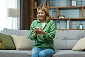 Happy senior woman sitting on sofa at home and using phone. Chats, reads messages, plays online games