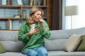 An attack of asthma, allergies at home. An elderly woman is sitting on the sofa, holding her chest and coughing, feeling severe pain, suffocating
