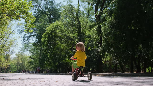 Blond little kid rides tricycle and looks around in park