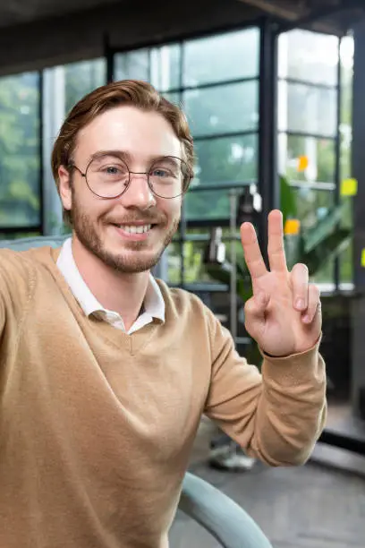 Photo of Close-up photo. Portrait of professional smart male programmer hacker taking selfie with hand, showing victory gesture with fingers. Smiles into the phone camera