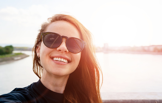 Young urban woman wearing sunglasses smiling and taking selfie against the river.