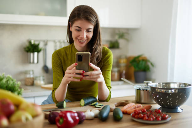 istock Young woman text messaging while cutting vegetables on cutting board in the kitchen 1487520187