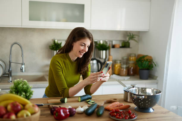 istock Young woman text messaging while cutting vegetables on cutting board in the kitchen 1487520247