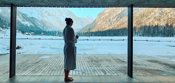A woman standing in a sauna relaxation area with a beautiful view of the winter mountains. Concept of winter SPA and recreation on nature.
