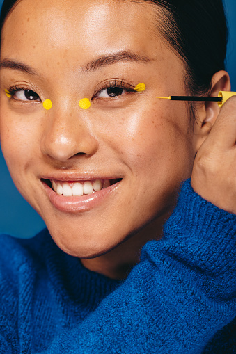 Asian female applying eyeliner on her face, creating four yellow dots. Beautiful young woman smiling and looking at the camera as she does a trendy makeup technique with vibrant eye makeup.