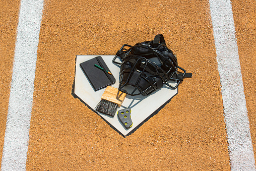 An umpire's equipment sitting on home plate with hints of the white lines of the batter's box. Their equipment consists of a rule book, note pad, pencil, brush, indicator for balls and strikes and a face mask.