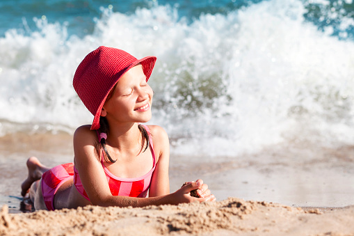 Happy Child Girl in Red Laying on Sunny Beach on Sea Wave Background. Smiling Teen Enjoying Sea,  Tanning on Sandy Beach. Happy Sea Vacation.