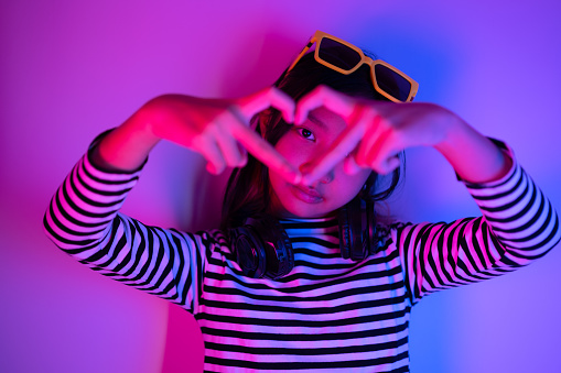 Portrait of an Asian girl wearing a black and white striped shirt and yellow sunglasses headband posing in a heart-shaped hand gesture with blue and red neon lights.