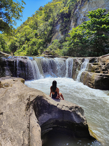 Close-up rear view shot of Asian woman sitting on the rock, watching a waterfall scenic view in Sumba island.