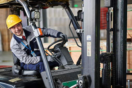 Side view of one asian warehouse worker driving forklift in distribution warehouse.