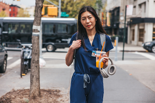 Young Asian (Korean) nurse commuting to work. Walking in New York city street texting on mobile