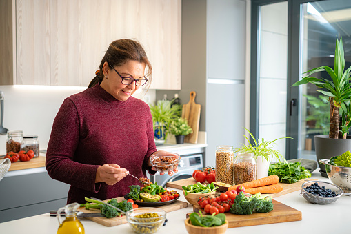 Portrait of a mature Hispanic woman preparing healthy vegan salad plate in domestic kitchen. Healthy eating concept. High resolution 42Mp indoors digital capture taken with SONY A7rII and Zeiss Batis 40mm F2.0 CF lens