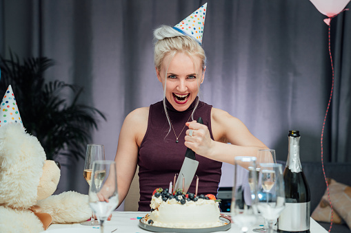 Attractive crazy laughing birthday woman with bread-knife planning to cut her birthday cake while celebrating birthday at home alone. Selective focus.