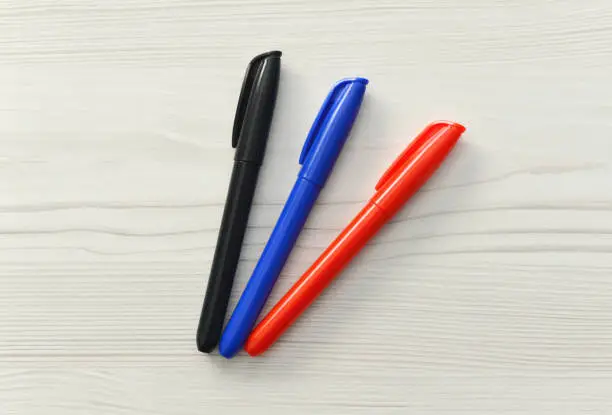 Closeup of three generic permanent markers in black, blue and red on a whitewashed wood background.