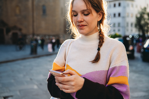 A woman is texting with a smart phone in the city at sunset. She's exploring San Lorenzo district in Florence, Italy.