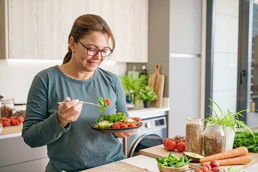 Portrait of a mature Hispanic woman eating healthy vegan salad plate in domestic kitchen. Healthy eating concept. High resolution 42Mp indoors digital capture taken with SONY A7rII and Zeiss Batis 40mm F2.0 CF lens