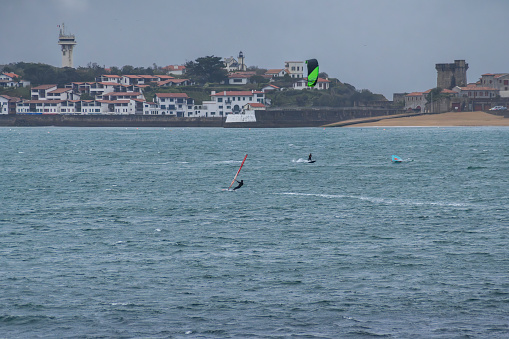 sailing and windsurfing and kitesurfing among the waves a day of bad weather with the sea somewhat choppy and the sky dark, with wind, in the bay of san juan de luz, the beach and the bay of sokoa, and the castle and the beach