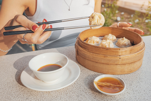 Girl with chopsticks eating delicious, traditional Chinese delicacy, Xiao long bao dumplings, or baozi buns presented in a bamboo plate with soy sauce for an authentic touch.