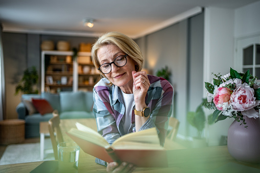 The photo shows a mature woman in her comfortable apartment, sitting in the kitchen area of her apartment and indulging in her favorite book. The gentle light of the morning sun comes through the window, the painting exudes peace and tranquility, depicting the daily routine of a woman who cherishes her quiet moments of solitude and the simple pleasures of reading.