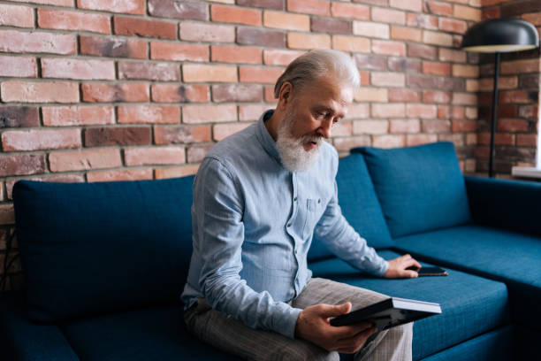 střední záběr nešťastného zamyšleného šedovlasého zralého muže při pohledu na rodinný portrét držícího fotorámeček sedícího sám doma na gauči. koncept nostalgie - štětkotvaré fotky - stock snímky, obrázky a fotky