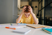 Portrait of exhausted pupil boy tired from studying holding head head with hands sitting at desk with paper copybook, looking down.