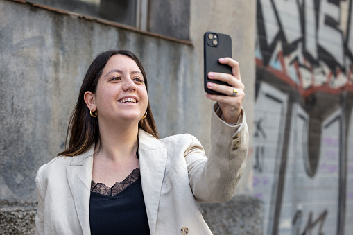 Young woman, generation z, in urban setting in Barcelona using her mobile phone. She has long brown hair and no glasses, wearing a beige and black shirt. She is looking friendly and is smiling. She is in front of an industrial building. Negative space for typo and part of a series.