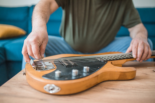 Unrecognizable male guitarist fixing bass guitar while sitting on sofa in living room