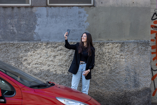 Young woman, generation Z, in urban setting in Barcelona using her mobile phone with red car in foreground. She is wearing blue jeans and black jacket. Her hair is long and straight. She is using her mobile phone while waiting behind a red car which is parked in the street.