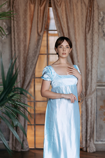 A young beautiful girl in a blue Regency dress stands at a wooden window in the estate. The classic artistic image of Russian literature of the 19th century, outdated.