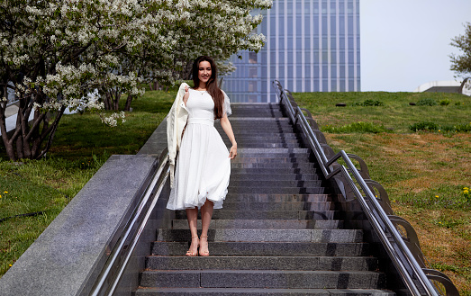girl in a white dress stands on the stairs in a big city next to flowering trees in spring
