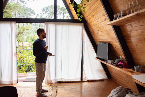 man standing with remote control turning on tv and glass door with nature in the background