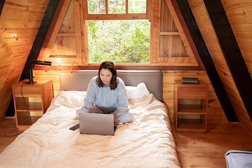 woman sitting with notebook on bed wearing winter clothes in log cabin