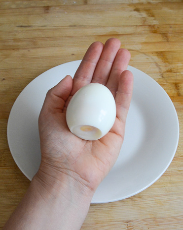 A caucasian hand holds a hard-boiled hen's egg above a white plate and wooden table background