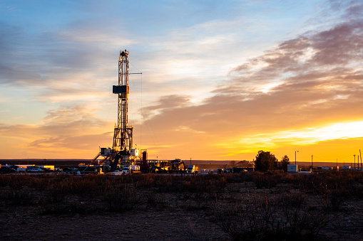 This photo depicts an oil drilling platform in New Mexico at sunrise. The golden light of the sun illuminates the metal structure, creating a vivid contrast with the dark shadows of the surrounding landscape.