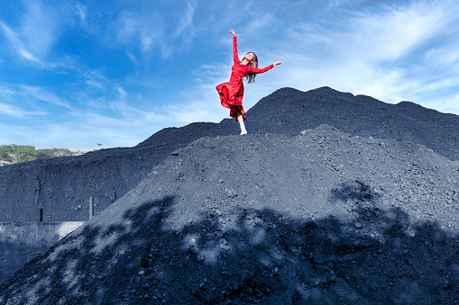 Full body of female with black flying hair in red dress raising leg while dancing on pile of coal against blue sky concept of uniqueness and sticking out from crowd