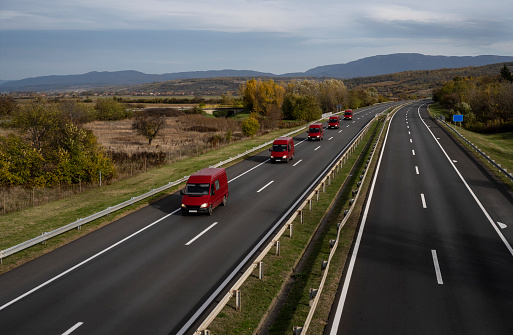 Five Red minivans are moving in a convoy on the highway. Red delivery van on the highway. Red modern delivery small shipment cargo courier van moving fast on motorway road to city urban suburb.