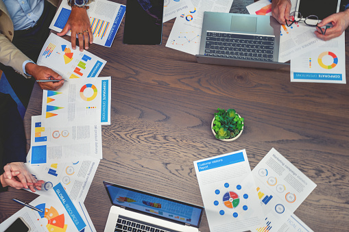 Paperwork and computers on a board room table at a business meeting presentation or seminar. The documents have financial or marketing figures, graphs and charts on them. People are pointing at the data. High angle view