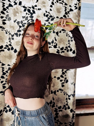 Cute and smiling red headed teen girl, at home, posing with a big red flower in front of her flowered covered curtains at home.