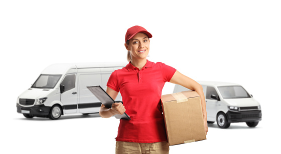 Female delivery worker holding a clipboard and a box in front of transport vans isolated on white background